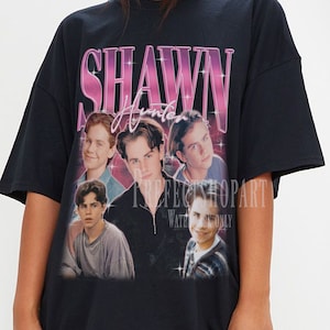 Könnte beinhalten: Schwarzes Oversize-T-Shirt mit einem grafischen Druck einer Gruppe junger Männer. Der Druck zeigt den Text "SHAWN Hunter" in rosa und weißen Buchstaben.