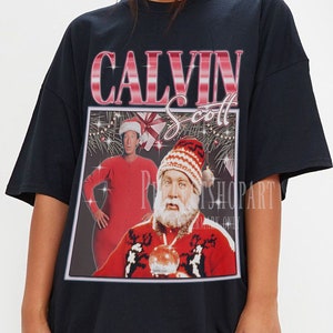 May include: Black oversized t-shirt featuring a graphic print of Calvin Scott dressed as Santa Claus. The graphic depicts a red and white Christmas sweater with a reindeer design and a red Santa hat.