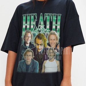 Könnte beinhalten: Schwarzes Oversize-T-Shirt mit einem Grafikdruck von Heath Ledger als Joker aus dem Film "The Dark Knight". Die Grafik enthält mehrere Bilder von Ledger in verschiedenen Szenen aus dem Film. Der Text "HEATH Ledger" ist in Grün und Weiß über den Bildern gedruckt.