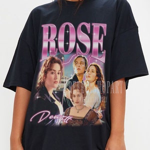 Könnte beinhalten: Schwarzes Oversize-T-Shirt mit einem Grafikdruck einer Szene aus dem Film Titanic. Der Druck zeigt die Figuren Rose und Jack, und der Text "ROSE" in Pink mit einem Glitzer-Effekt. Der Text "DeWitt" ist ebenfalls in Pink unter dem Bild gedruckt.
