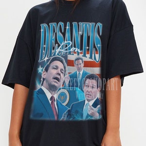 May include: Black oversized t-shirt with a vintage-style graphic of Ron DeSantis, a politician, with the text "Desantis" and "Ron" in blue. The graphic also includes an American flag and the text "Keep Florida Great" and "Water is Life" in smaller font.