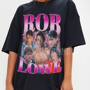 May include: Black t-shirt with a retro graphic of Rob Lowe and other actors. The design features the name "ROB LOWE" in a pink gradient font. The shirt has a relaxed, oversized fit.