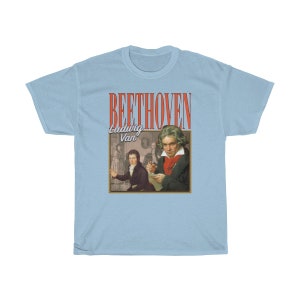 Pu&ograve; includere: T-shirt blu chiaro con una grafica in stile vintage di Ludwig van Beethoven. La grafica presenta un ritratto di Beethoven e il testo "BEETHOVEN Ludwig Van" in rosso e bianco.