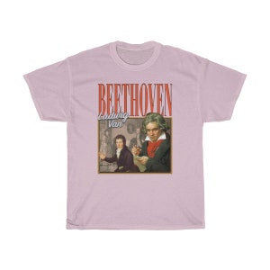 Pu&ograve; includere: Una t-shirt rosa chiaro con una grafica in stile vintage di Ludwig van Beethoven. La grafica presenta un ritratto di Beethoven e il testo "BEETHOVEN Ludwig Van" in rosso e bianco.