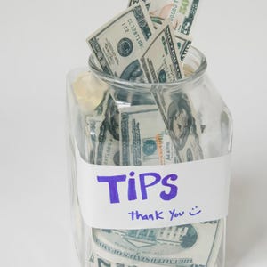 Könnte beinhalten: Ein klares Glasgefäß gefüllt mit US-Dollar-Scheinen. Ein weißes Etikett um das Glas trägt in lila Marker die Aufschrift "TIPS", darunter "thank you" und ein Smiley. Das Glas ist überfüllt mit verschiedenen Banknoten.