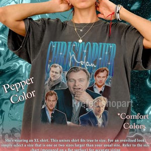 May include: Pepper-colored unisex t-shirt featuring a graphic of Christopher Nolan's name in blue and white, with images of the director and actors. The shirt has the words "Comfort Colors" printed on it.