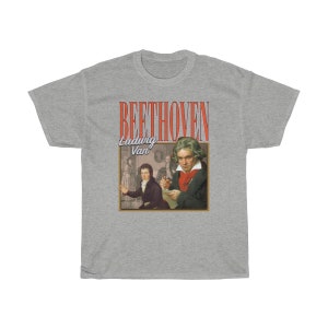 Pu&ograve; includere: Una t-shirt grigia con una grafica in stile vintage di Ludwig van Beethoven. La grafica presenta un ritratto di Beethoven e il testo "BEETHOVEN Ludwig Van" in rosso e bianco.
