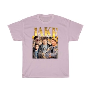May include: A light pink t-shirt with a gold graphic of Jake Peralta from the TV show Brooklyn Nine-Nine. The graphic includes the text "JAKE PERALTA" and features multiple images of Jake Peralta in different poses.