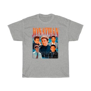 Peut inclure: T-shirt gris avec un graphique de style vintage repr&eacute;sentant un groupe d'hommes avec le texte "Jonathan Bailey" dans une police rouge et orange.