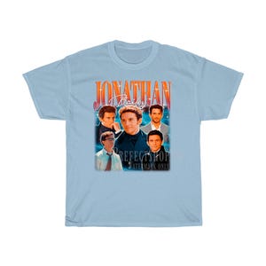 Peut inclure: T-shirt bleu clair avec un graphique de style vintage repr&eacute;sentant un groupe d'hommes avec le texte "Jonathan Bailey" dans une police rouge et orange.