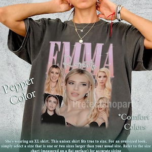 May include: A charcoal gray short-sleeve t-shirt featuring a graphic print of the name Emma Roberts in pink lettering above a collage of five different photographs of the actor Emma Roberts. The shirt has a crew neckline and a relaxed fit.