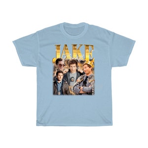 May include: Light blue t-shirt with a gold graphic of Jake Peralta from the TV show Brooklyn Nine-Nine. The graphic features Jake in different poses, including wearing a crown and sunglasses. The text "JAKE PERALTA" is written in gold letters above the graphic.