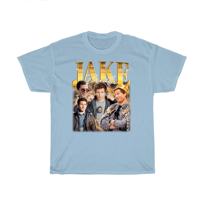 May include: Light blue t-shirt with a gold graphic of Jake Peralta from the TV show Brooklyn Nine-Nine. The graphic features Jake in different poses, including wearing a crown and sunglasses. The text "JAKE PERALTA" is written in gold letters above the graphic.