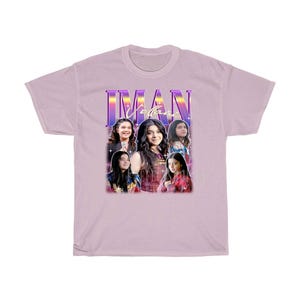May include: A light purple t-shirt with a retro-style graphic featuring a group of four people. The graphic includes the text "IMAN" in a purple and blue gradient. The people in the graphic are all women with dark hair and are wearing colorful clothing.