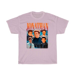 Peut inclure: Un t-shirt rose p&acirc;le avec un graphique de style vintage repr&eacute;sentant un groupe d'hommes avec le texte "Jonathan Bailey" en caract&egrave;res orange gras.