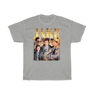 May include: A gray t-shirt with a gold graphic of Jake Peralta from the TV show Brooklyn Nine-Nine. The graphic includes the text "JAKE" and "Peralta" in gold lettering.