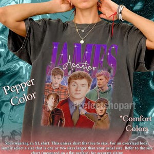 May include: A dark gray unisex t-shirt featuring a graphic of James Acaster with a purple and white design. The shirt has the text "JAMES Acaster" in a retro style. The shirt is labeled "Pepper Color" and "Comfort Colors".