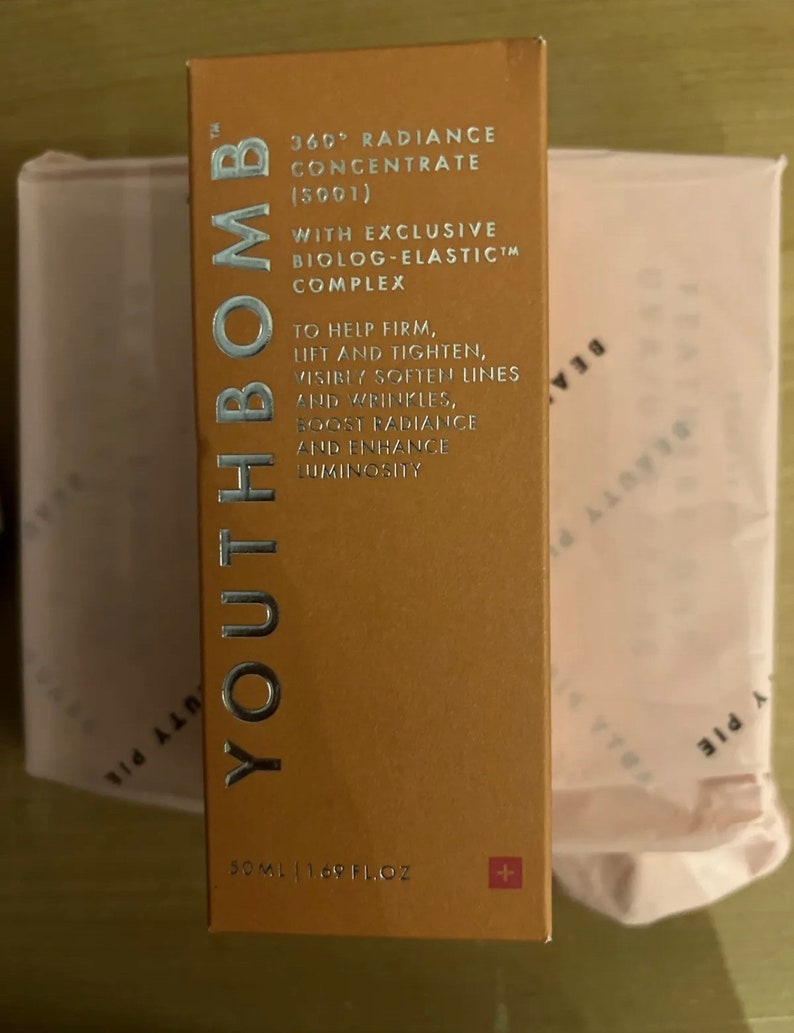 May include: A gold box with the text "Youth Bomb" in silver lettering. The box is labeled "360&deg; Radiance Concentrate (5001)" and "With Exclusive Biolog-Elastic Complex". The box also says "To help firm, lift and tighten, visibly soften lines and wrinkles, boost radiance and enhance luminosity". The box is 50 ml (1.69 fl oz) and has a small Swiss flag on the bottom right corner.