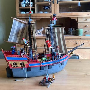 Peut inclure: Un modèle de navire pirate détaillé avec des accents rouges, bleus et gris. Le navire comprend plusieurs mâts, voiles et figurines de pirates. Un petit canot avec un pirate est devant le navire. Un drapeau noir avec une tête de mort est visible.
