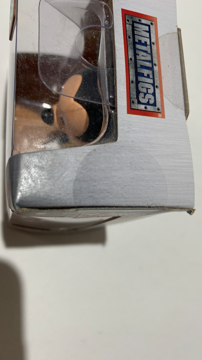 May include: A partially visible product package with the text "METALFIGS" in a rectangular frame. The package is silver with a clear plastic window. The product inside is partially visible.