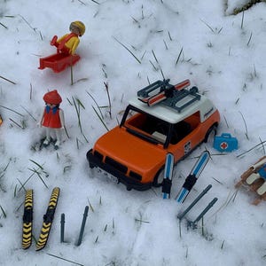 Op de afbeelding: Een set met speelgoedfiguren en accessoires voor een winterscène. De set bevat een rode en witte speelgoedauto met ski's op het dak, twee paar ski's, een rode slee en drie speelgoedpoppetjes. De poppetjes dragen winterkleding en zijn geposeerd in een besneeuwde omgeving.