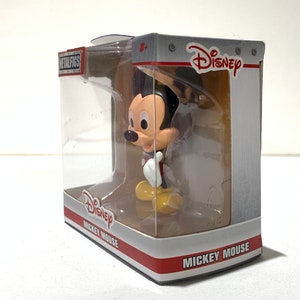 May include: A Mickey Mouse figurine in a clear plastic display case. The figure is in a classic pose, wearing red shorts, yellow shoes, and white gloves. The packaging features the Disney logo and the words "Mickey Mouse."