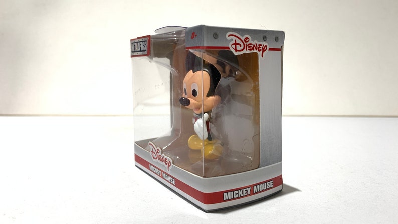 May include: A Mickey Mouse figurine in a clear plastic display case. The figure is in a classic pose, wearing red shorts, yellow shoes, and white gloves. The packaging features the Disney logo and the words "Mickey Mouse."