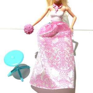May include: A blonde doll wearing a white strapless dress with pink floral patterns and a pink tiara. The doll is holding a pink bouquet and is standing on a blue stool.