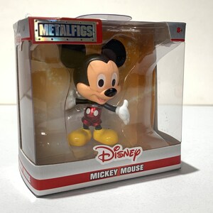 May include: Mickey Mouse figure in a clear plastic display case. The figure has a black head and ears, a tan face, white gloves, red shorts, and yellow shoes. The packaging includes the "METALFIGS" logo and "Disney MICKEY MOUSE" text.