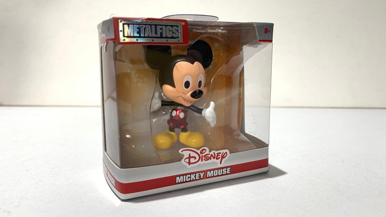 May include: Mickey Mouse figure in a clear plastic display case. The figure has a black head and ears, a tan face, white gloves, red shorts, and yellow shoes. The packaging includes the "METALFIGS" logo and "Disney MICKEY MOUSE" text.