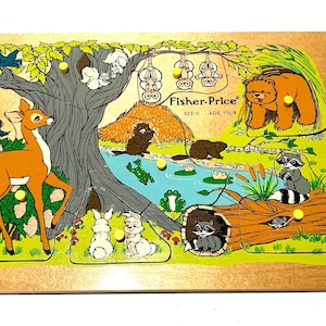 Op de afbeelding: Houten Fisher-Price puzzel met een bosscène met een hert, een beer, wasberen en andere dieren. De puzzel heeft een boom, een vijver en een boomstam. De tekst "Fisher-Price" en "523-E AGE 1 1/2-4" zijn zichtbaar.