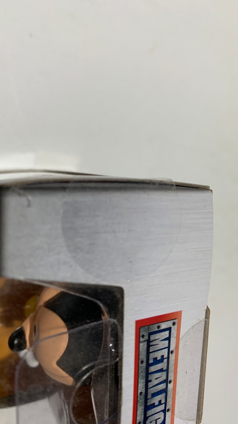May include: A partially visible action figure in a clear plastic case. The figure has a black and tan color scheme. The packaging has a white background with the words "METALFIG" in a red and silver design.