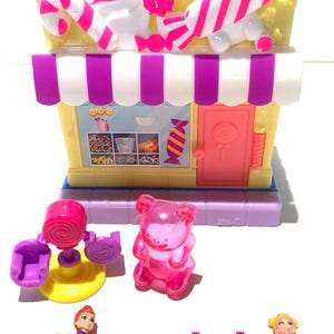 Op de afbeelding: Een roze en witte snoepwinkel speelset met een roze gummybeer, twee poppen en een draaiende snoepstokstand. De winkel heeft een paars en wit gestreept luifel en een roze deur.