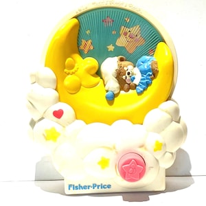 Könnte beinhalten: Vintage Fisher-Price Musik-Nachtlicht mit gelbem Mond, weißen Wolken und einem schlafenden Teddybär. Das Nachtlicht hat einen türkisfarbenen Hintergrund mit Sternen und einen rosa Knopf. Auf der Basis des Nachtlichts befindet sich das Fisher-Price-Logo.