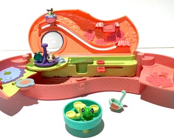 May include: A coral-colored, open playset with a slide, a pond, and a rotating platform. The playset includes a small, round mirror, a green and yellow counter, and several small animal figurines. A teal bowl with green frog figurines and a small ladle are also included.