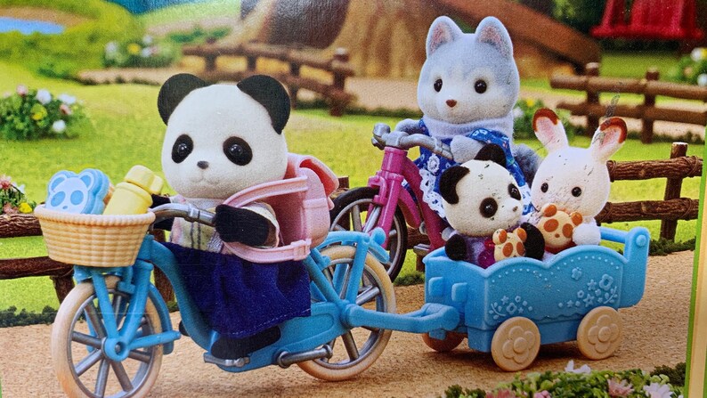 May include: A panda figure with a pink backpack rides a blue bicycle with a front basket containing a yellow bottle and a blue bear-shaped item. A blue trailer attached to the bike carries a small panda figure and a white rabbit figure.