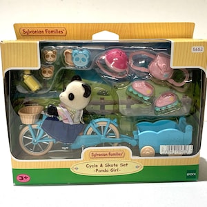 May include: A Sylvanian Families Cycle & Skate Set featuring a panda figure on a light blue bicycle with a matching trailer. The set includes a pink helmet, a pink backpack, two pairs of roller skates, a yellow water bottle, and small accessories.