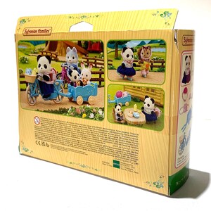 May include: A Sylvanian Families toy box featuring illustrations of panda and rabbit figures with a blue bicycle and trailer. The packaging displays various scenes of the animal characters in a miniature outdoor setting with printed text.