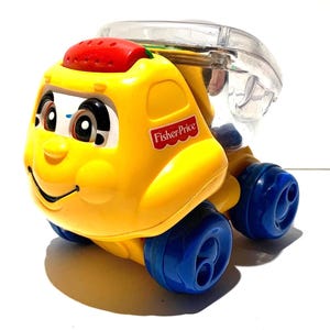 Könnte beinhalten: Ein gelber Fisher-Price Spielzeug-LKW mit einem lächelnden Gesicht, roter Oberseite und blauen Rädern. Das Spielzeug hat einen durchsichtigen, abgerundeten Behälter oben. Das Fisher-Price-Logo ist auf der Seite des LKWs sichtbar.