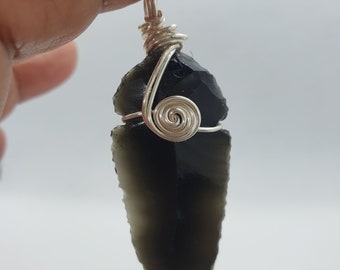 Pode incluir: Um pingente de ponta de flecha de obsidiana preta envolto em fio de prata. O fio é torcido em um design espiral na parte superior do pingente.