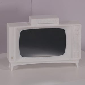 Könnte beinhalten: Ein weißer Fernseher im Vintage-Stil mit einem schwarzen Bildschirm und einem weißen rechteckigen Kasten oben. Der Fernseher steht auf einer weißen Oberfläche.