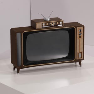 Pode incluir: Uma televisão de estilo vintage com um pequeno rádio em cima. A TV tem uma moldura de madeira castanha escura com detalhes dourados e um ecrã preto. O rádio também é castanho com detalhes prateados e uma antena. O conjunto está sobre uma superfície branca.