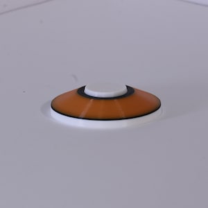 May include: A small, orange and black circular object with a white top. The object is made of plastic and has a smooth surface.