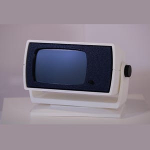 Op de afbeelding: Een vintage zwart-witte televisie met een klein scherm. De tv heeft een rechthoekige vorm met een donker scherm en een zwarte knop aan de zijkant. Hij is op een witte standaard gemonteerd.