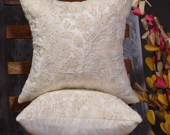 May include: Two white embroidered throw pillows with a floral design. The pillows are stacked on top of each other and are sitting on a wooden chair.