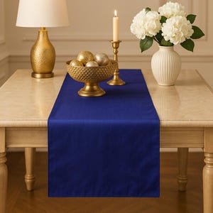 May include: A navy blue table runner is centered on a light-colored wooden table. A gold-colored lamp, a decorative bowl with gold spheres, a candle holder with a lit candle, and a white vase with white flowers are arranged on the table.