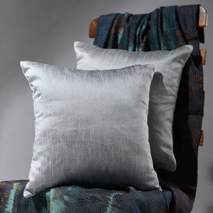 May include: Two silver gray throw pillows with a subtle textured pattern are arranged on a dark blue and green patterned throw blanket.