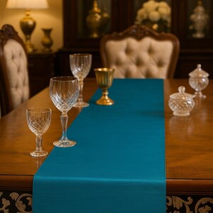 May include: A teal blue table runner with a fringed edge is centered on a wooden table. Crystal wine glasses and a gold goblet are arranged on the runner. A crystal sugar bowl sits to the right.