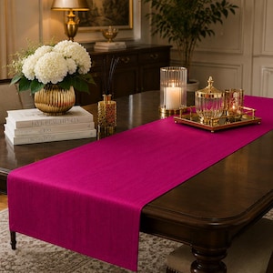 May include: A dining table set with a vibrant fuchsia table runner. The table is adorned with a gold vase of white flowers, books, candles, and decorative glass pieces. The room has a classic, elegant aesthetic.