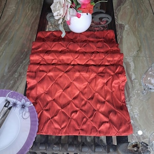 May include: A red quilted table runner with a diamond pattern. The runner is folded in half and placed on a glass table.